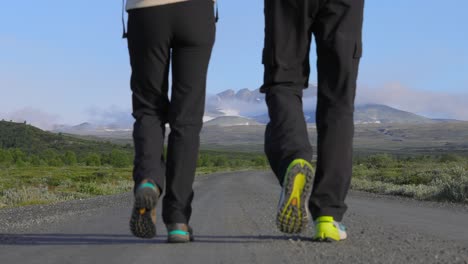 Pareja-Caminando-Con-Mochilas-Por-Una-Carretera-Con-Montañas-Al-Fondo.-El-Parque-Nacional-Dovrefjell-Sunndalsfjella-Es-Un-Parque-Nacional-En-Noruega.-Hermoso-Paisaje-Natural-De-Noruega.